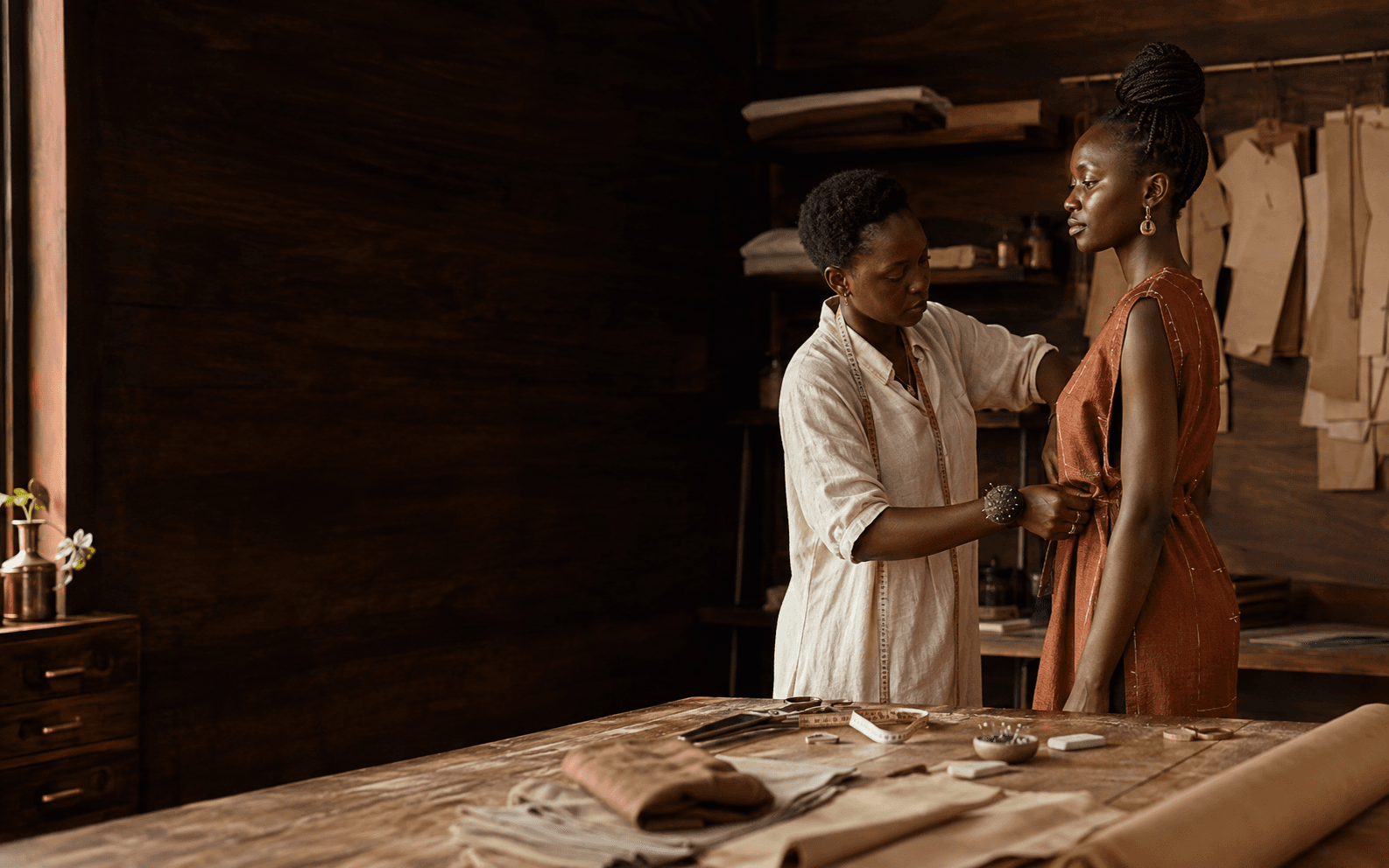 A Black fashion designer carefully fitting a client in a warm, working atelier